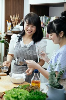 一位快乐的亚洲成年女士戴着围裙，和十几岁的女儿一起站在厨房的木桌旁，边做饭边讨论食谱细节