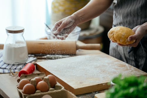 Crop faceless female baker in apron sprinkling wheat flour on cutting board to roll out fresh dough in modern kitchen
