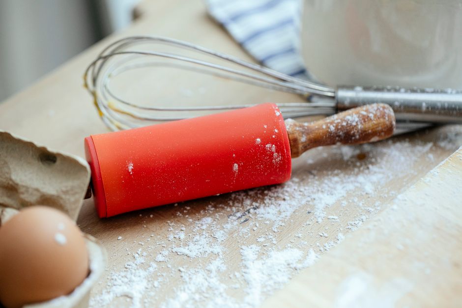Wooden table with rolling pin and whisk and eggs near chopping board covered with flour in kitchen