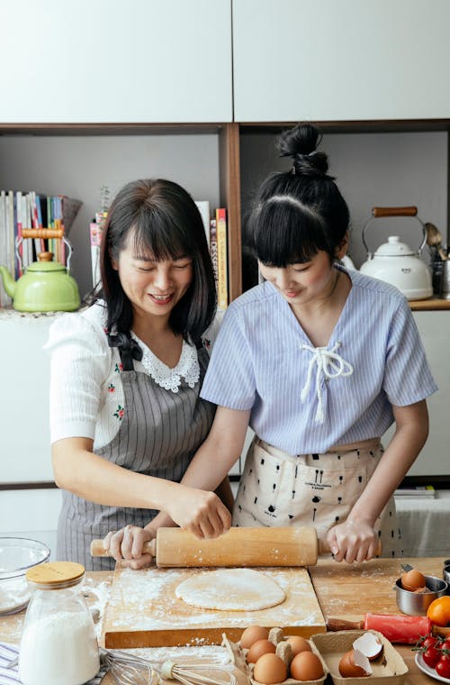 Smiling mother helping daughter making dough with rolling pin for pastry in kitchen