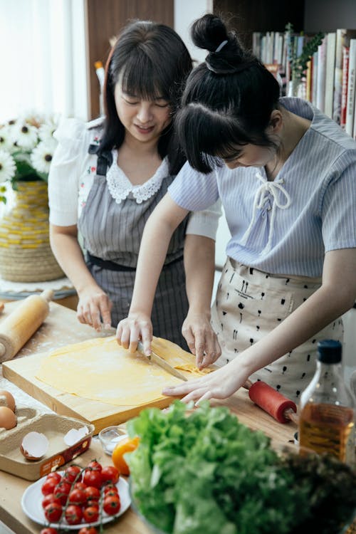 Free High angle of Asian women in aprons cutting dough for pasta on table with vegetables eggs and cooking ingredients Stock Photo