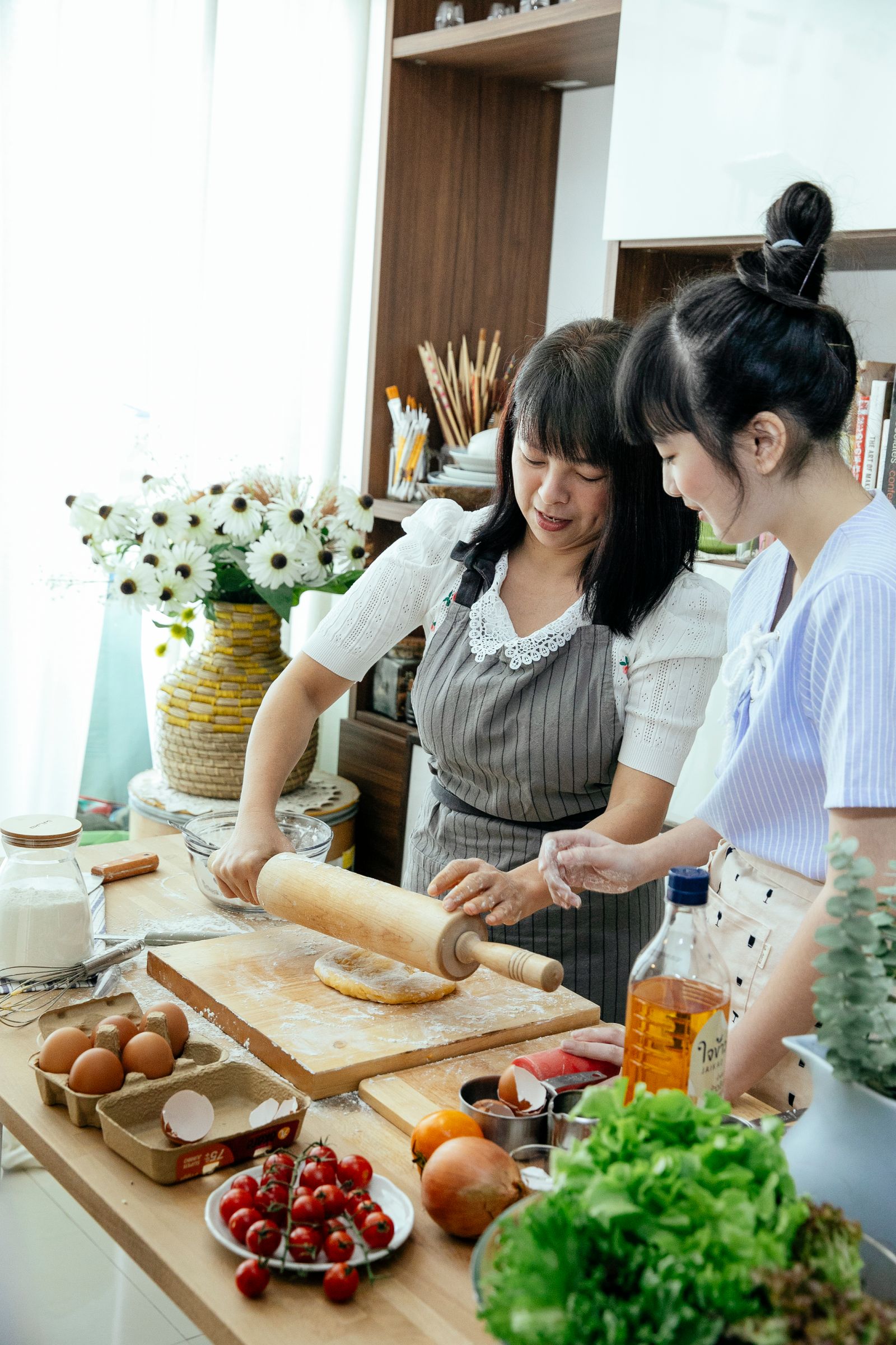 A woman in an apron and a younger woman rolling dough together at a kitchen table
