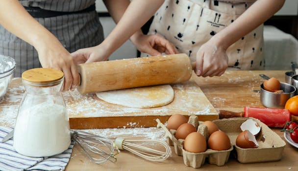 Unrecognizable women rolling dough together on board on table with jar with flour carton with eggs and whisks