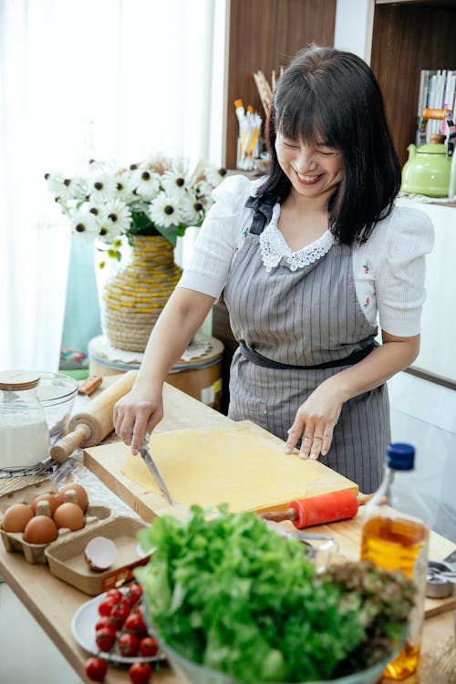 Free Cheerful Asian female in apron cutting rolled dough on chopping board on table with cooking ingredients in kitchen Stock Photo