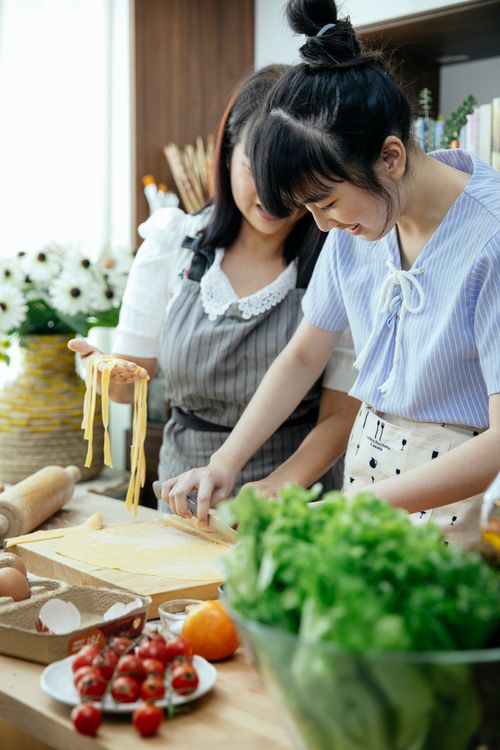 Free Positive Asian women in aprons making homemade long egg noodles with dough on table with cooking ingredients in kitchen Stock Photo