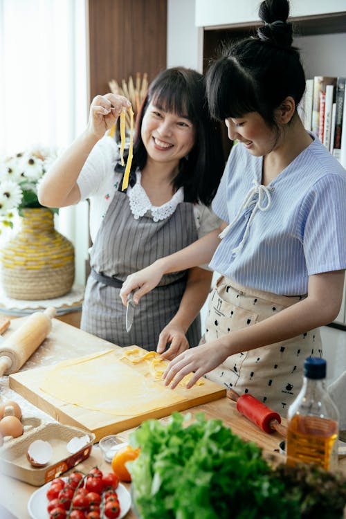 Free Cheerful Asian women in aprons making together homemade long noodles on table with chopping board and cooking ingredients in kitchen Stock Photo