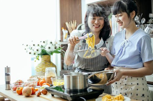 Free Cheerful Asian teen girl with middle aged mother draining boiled pasta in stainless bowl cooking in team Stock Photo