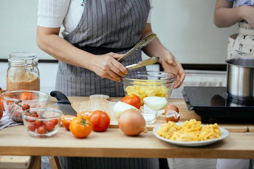 Free Faceless women in aprons cooking delicious pasta with fresh tomatoes standing at counter in modern kitchen Stock Photo