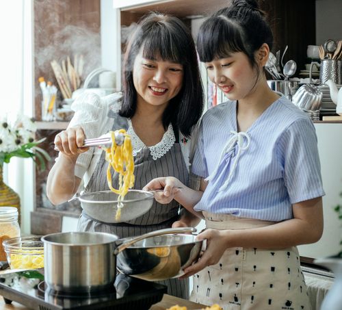 Free Smiling ethnic teenage with mother serving hot cooked pasta from saucepan into bowl Stock Photo