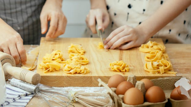 Faceless females in aprons at wooden table making homemade pasta cutting dough into strips