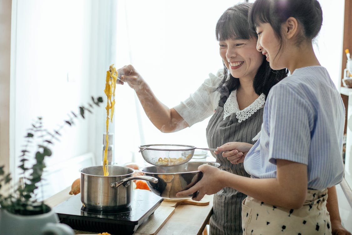 Free Side view of smiling Asian teenager with middle age mother serving hot boiled pasta in drainer Stock Photo