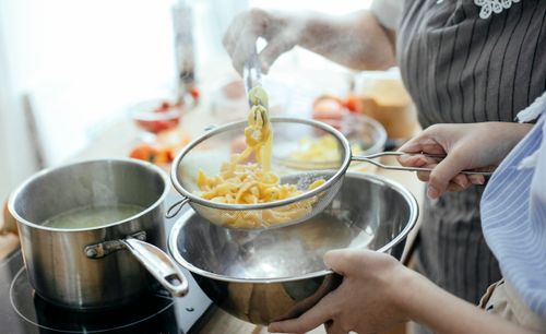 Free High angle of crop faceless female chefs putting noodles from saucepan with boiling water into strainer while standing at table with ingredients in kitchen Stock Photo
