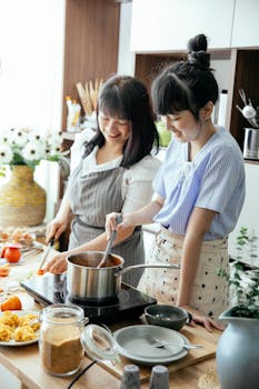 Cheerful Asian woman with young daughter smiling while cutting veggies and stirring sauce during lunch preparation together in kitchen