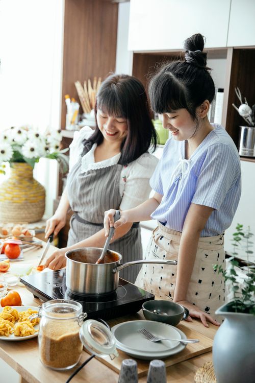 Free Cheerful Asian woman with young daughter smiling while cutting veggies and stirring sauce during lunch preparation together in kitchen Stock Photo