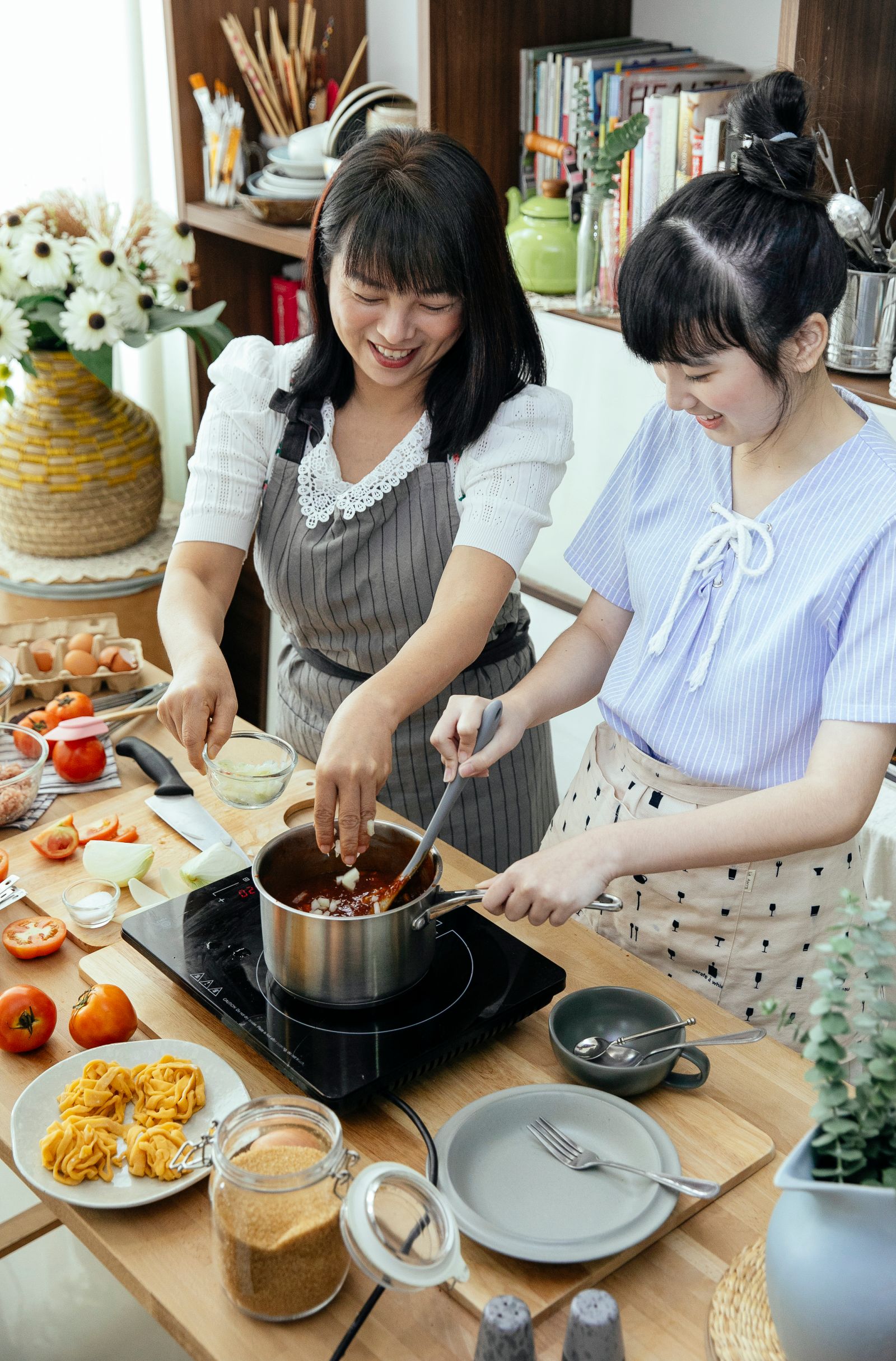 Happy Asian ladies standing near stove in aprons and stirring meal while cooking in light kitchen