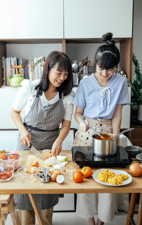 Positive Asian mother cutting vegetables while standing at table near daughter stirring food in pan while cooking together in kitchen