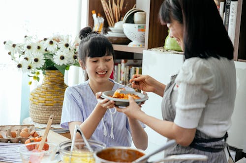 Free Delighted Asian female housewives in aprons mixing pasta with sauce in plate while cooking together in kitchen Stock Photo