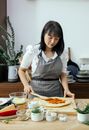Calm adult Asian housewife in apron spreading on raw pizza dough while cooking in kitchen