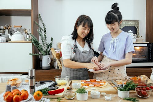 Happy smiling Asian mother and daughter in aprons putting ingredients on dough while cooking pizza together in kitchen