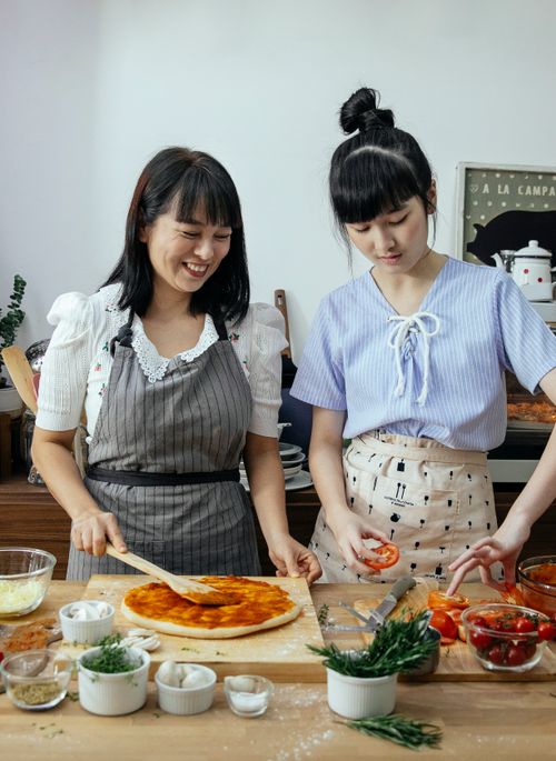Free Delighted Asian females in aprons putting tomatoes on dough with sauce while making pizza in kitchen Stock Photo