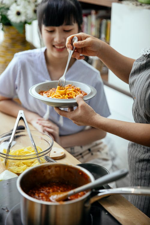 Free From above crop Asian women in aprons having homemade pasta with tomato sauce in kitchen Stock Photo