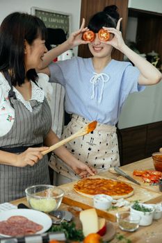 Laughing Asian mother and daughter in aprons cooking pizza together and having fun with tomatoes
