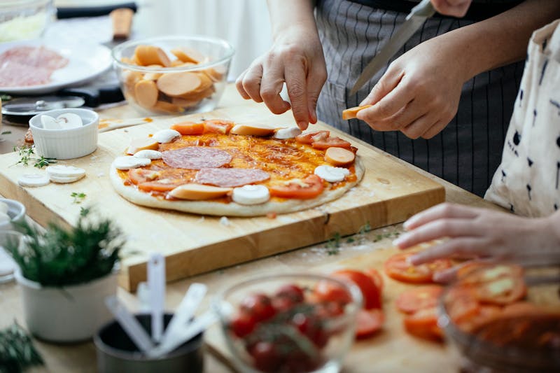 Crop anonymous person in apron adding sausages on pizza with salami and champignons while cooking in kitchen