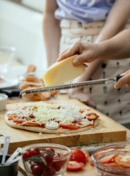 Crop unrecognizable person grating cheese on pizza with champignons and tomatoes while cooking in kitchen
