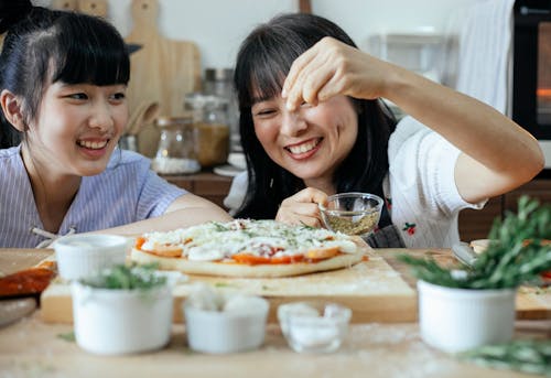 Happy Asian mother and daughter smiling while sprinkling seasoning yummy pizza while cooking together on cozy kitchen with various ingredients on table