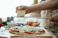 Side view of crop unrecognizable person grating piece of hard cheese on palatable homemade pizza in kitchen at daytime