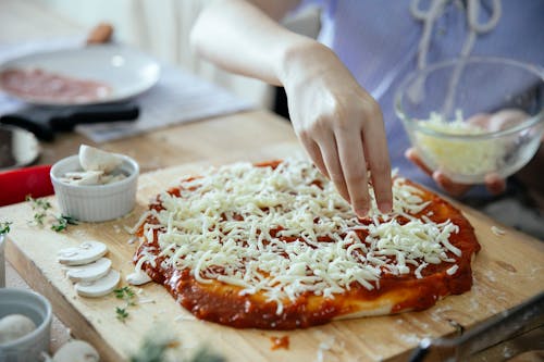 From above of crop anonymous female adding cheese from bowl on uncooked yummy pizza with tomato sauce  on wooden table in kitchen