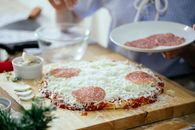 Crop anonymous person standing near table with uncooked homemade pizza and adding slices of salami while preparing in kitchen at daytime