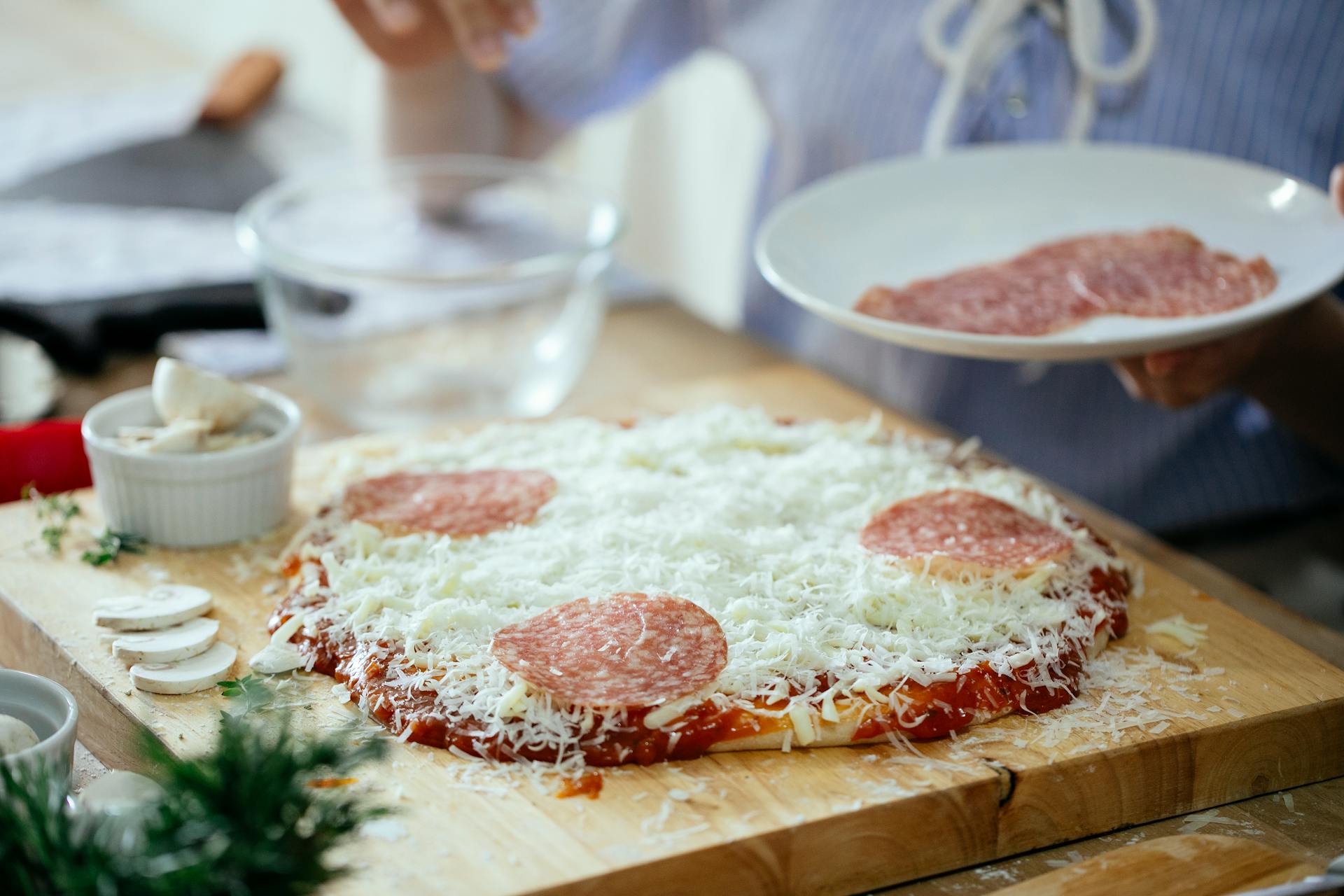 Crop anonymous person standing near table with uncooked homemade pizza and adding slices of salami while preparing in kitchen at daytime