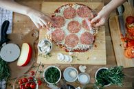Top view of crop unrecognizable people adding salami in homemade pizza placed on wooden table among various fresh ingredients while cooking together