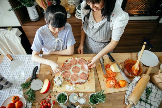 Overhead crop Asian mother and daughter cooking homemade pizza together on wooden table with various fresh ingredients in kitchen