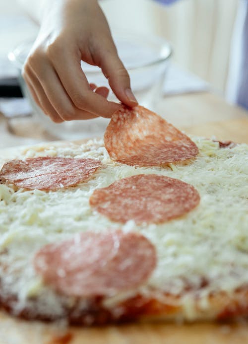 Crop unrecognizable person adding slice of salami on cheese layer while cooking homemade yummy pizza in kitchen