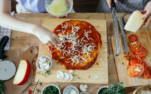 Free Overhead of crop unrecognizable women adding cheese from bowl in yummy pizza base with tomato sauce on wooden table among various fresh ingredients Stock Photo