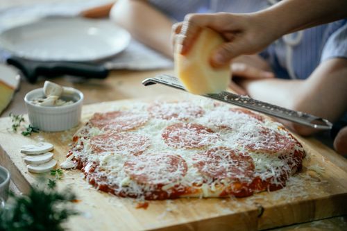 Free From above side view of unrecognizable crop person grating cheese on homemade yummy pizza placed on wooden table in kitchen Stock Photo