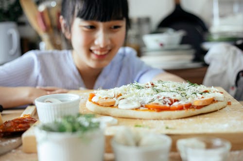 Crop Asian lady smiling and looking at homemade pizza placed on wooden table among various fresh ingredients and utensil in kitchen