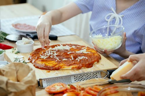 Free Side view of crop unrecognizable women sprinkling cheese from bowl on yummy homemade pizza while cooking together in kitchen Stock Photo