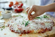 Crop unrecognizable person decorating yummy pizza with cheese cover by arugula on wooden table while cooking in light kitchen