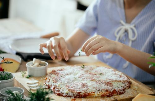 Free Unrecognizable woman sprinkling herbs on top of  homemade pizza sitting at table with cooking ingredients Stock Photo