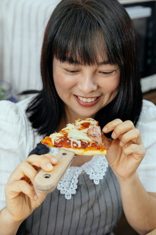 Free Cheerful Asian woman taking slice of cheesy and tomato sauce pizza from wooden spatula Stock Photo