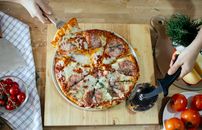 From above unrecognizable women taking slice of pizza with cheese and salami using spatula and cutting with pizza cutter