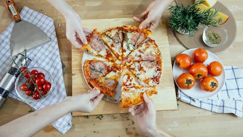 Free From above unrecognizable people taking slices of pizza with salami melted cheese and herbs from plate on kitchen table Stock Photo
