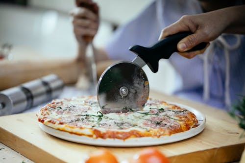 Free Unrecognizable person cutting pizza with melted cheese and herbs using pizza cutter in kitchen Stock Photo