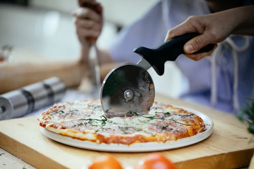 Unrecognizable person cutting pizza with melted cheese and herbs using pizza cutter in kitchen