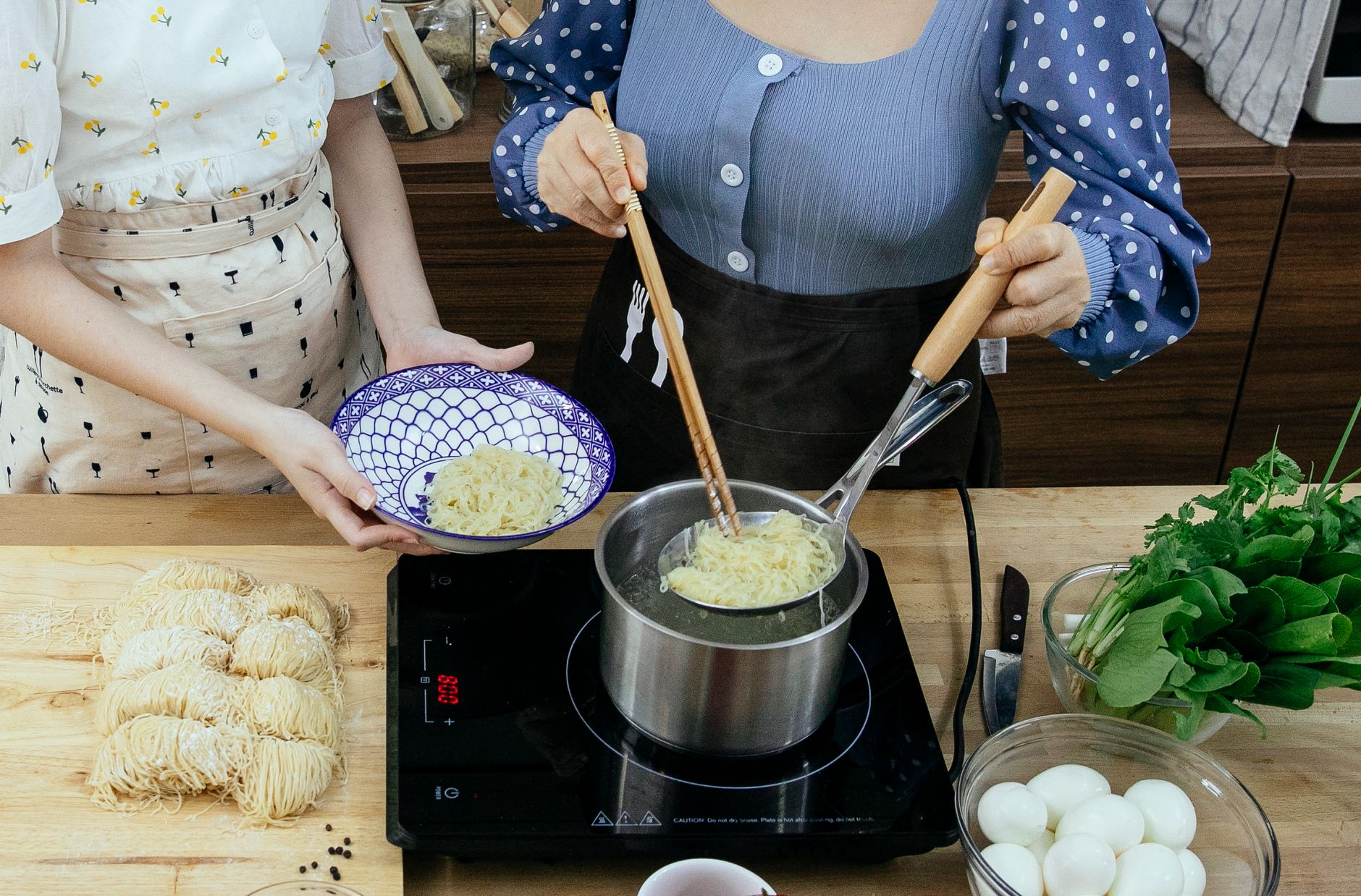 Unrecognizable women putting blanched noodles on plate while cooking Asian food on kitchen table with electric hob bowl with eggs and chopping board with raw noodles