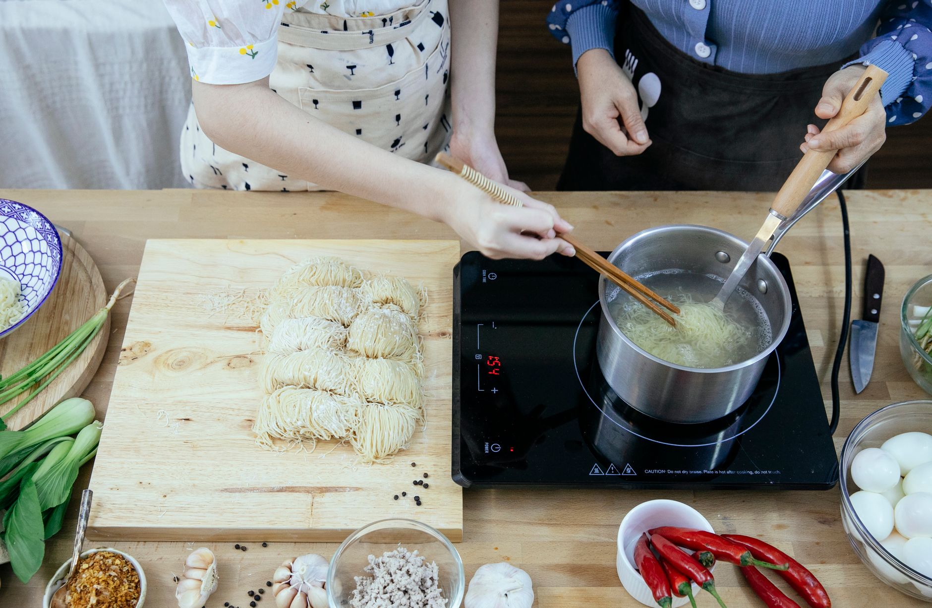 深鍋型小鍋煮麵防噴湯?三次實測揭曉真相 Crop anonymous housewives in aprons boiling noodles on tabletop stove near fresh ingredients in modern kitchen
