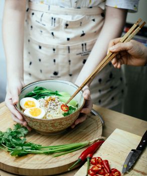 Crop anonymous females in aprons cooking together and decorating fresh Asian noodle soup with cut hot red pepper in light kitchen